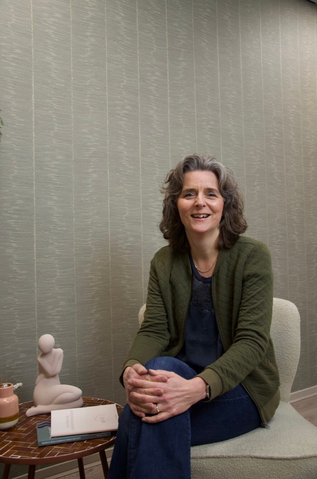 Nicole zit lachend in een fauteuil in haar praktijk, naast een beeldje en een boek
